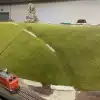 Foto einer Modelleisenbahnlandschaft: Grüner Hügel mit roter Dampflok, die von links ins Bild fährt